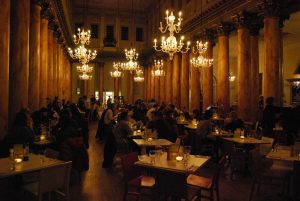 Photograph of ballroom style resturant venue with ornate columns and chandelier light fittings. People sit at tables in three rows up and down the floor  