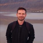 Photo of Kamil Karczewski from the waist up stood on a beach with cliffs in the background. he wears all black clothing and has short dark brown hair and a short dark brown beard. He is a white man in his early to mid 30s