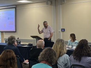 Photograph of the back of the heads of a group of adult people in casual business dress in an  off white painted seminar room looking at a white man with a beard in a short sleeve shirt stood up speaking with a lectern in front of him 